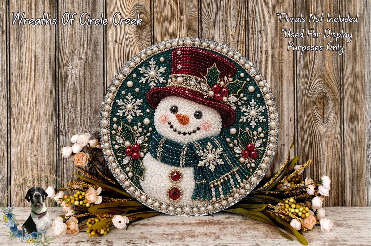 A snowman decoration with a red hat, green scarf, and beaded details, surrounded by a wreath of flowers and a wooden background.