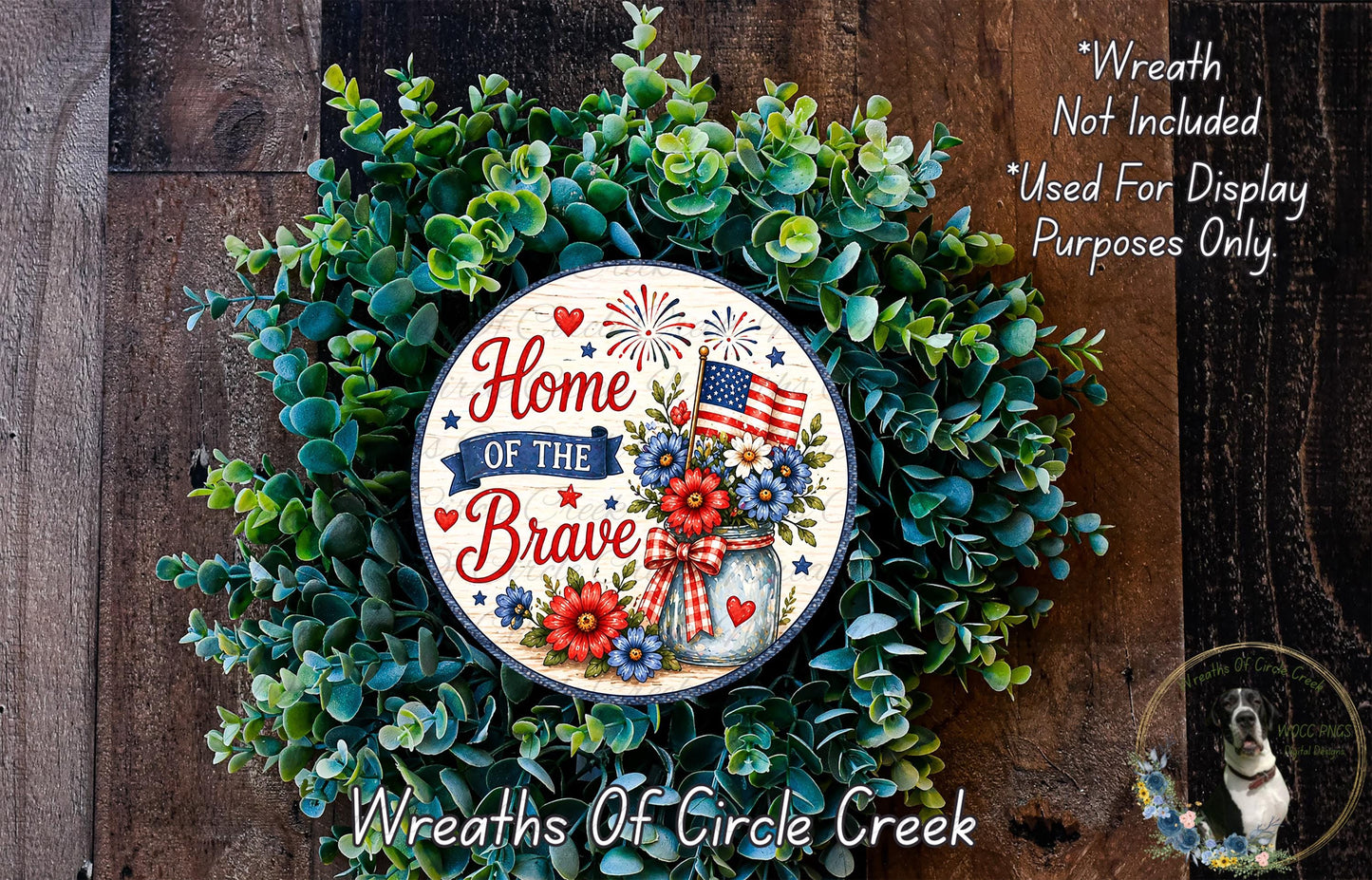 a wreath with the text "Home of the Brave" and an American flag, surrounded by green foliage. The wreath is displayed on a wooden surface, and there is a small dog in the bottom right corner of the image.