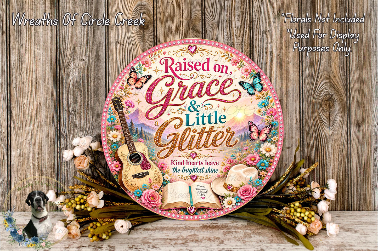 a decorative sign with a guitar, flowers, and butterflies, surrounded by a wreath. The sign has a pink and gold color scheme and displays the text "Raised on Grace & Little Glitter" and "Kind hearts leave the brightest shine".