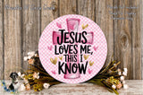 A pink and white sign with the text "JESUS LOVES ME THIS I KNOW" surrounded by flowers and greenery.