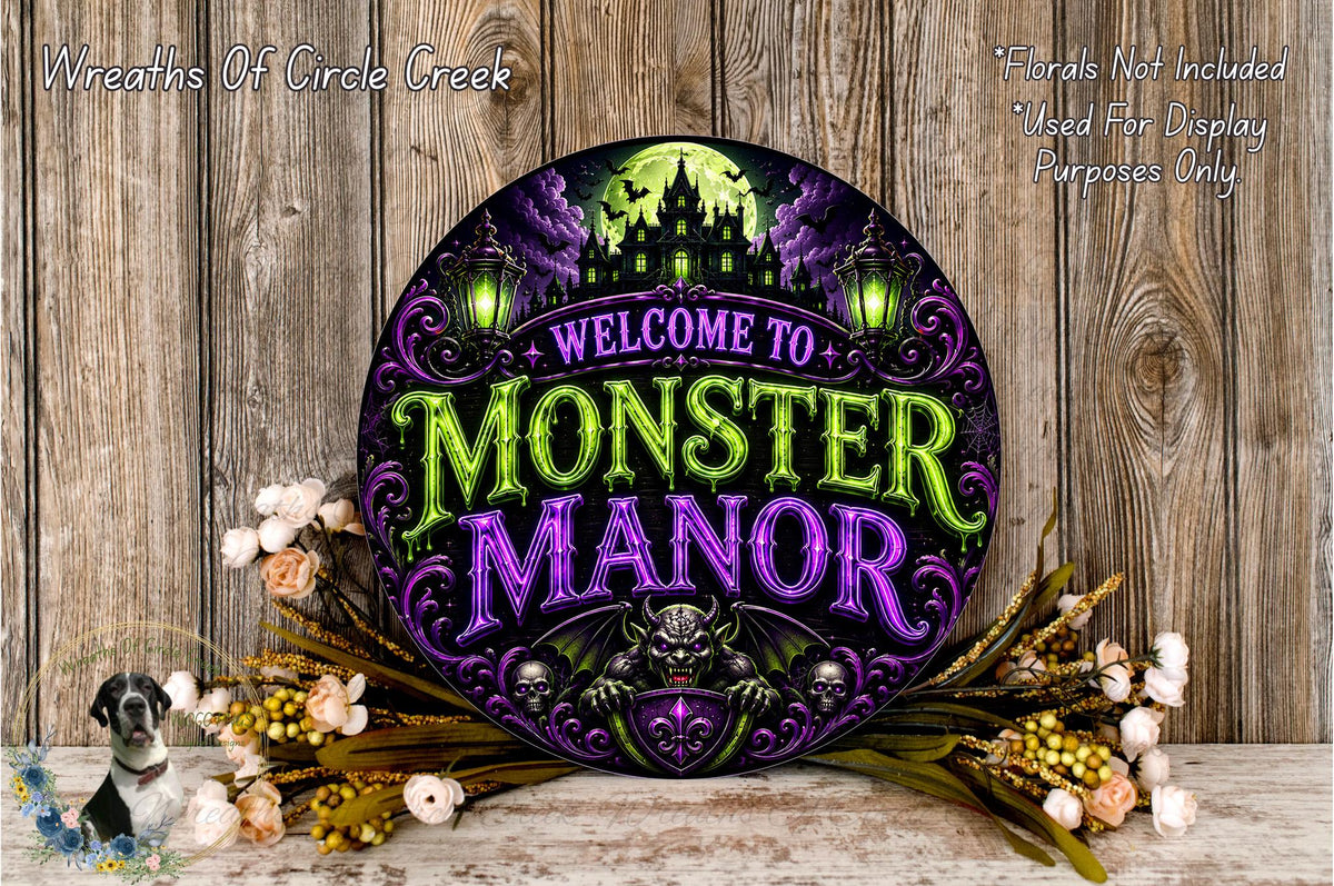 a round sign with a purple and green color scheme, displaying the text "Welcome to Monster Manor" in a decorative font. The sign is surrounded by flowers and placed on a wooden surface.