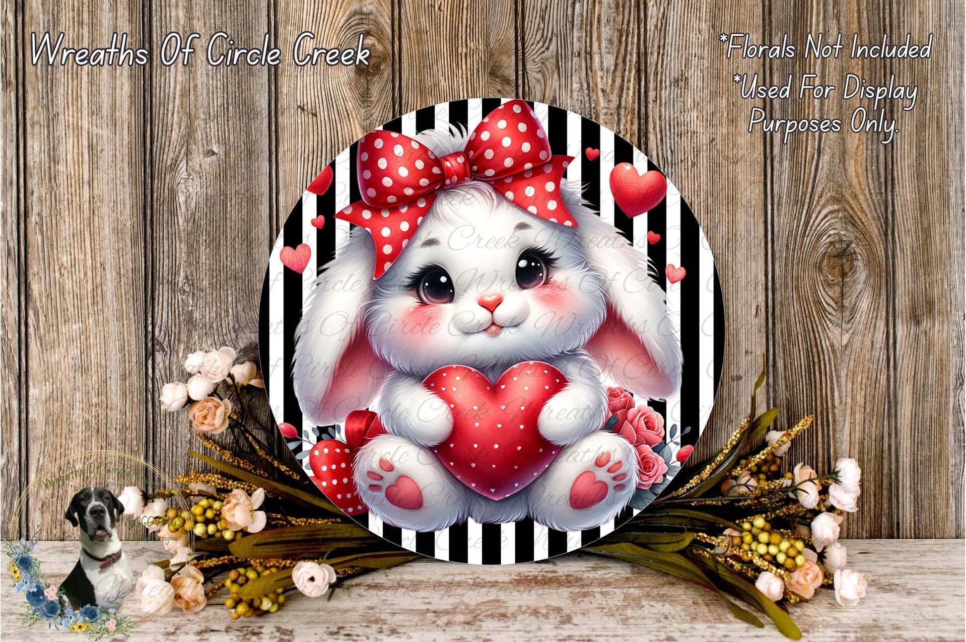A round, decorative wreath featuring a white bunny with a red bow and heart-shaped decorations, surrounded by flowers and greenery.