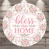 A round floral sign with the text "bless this home" in a decorative font, surrounded by pink roses and greenery.
