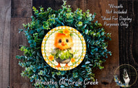 a wreath with a small, orange chicken as the central decoration, surrounded by a lush, green plant. The wreath is displayed on a wooden surface, and there is a small dog visible in the bottom right corner of the image.