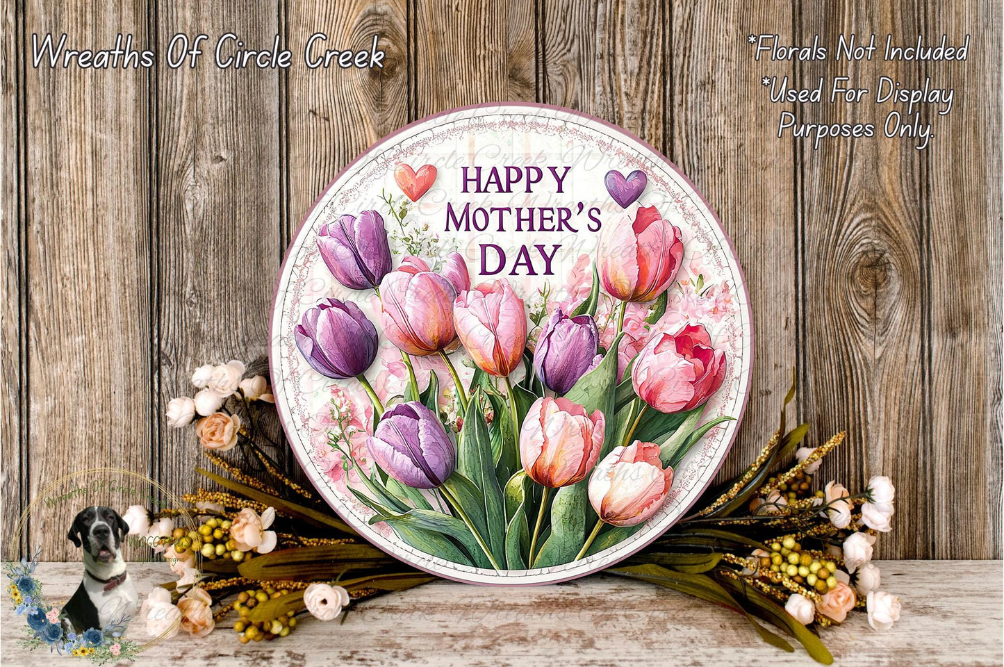A round floral wreath with a message that reads "Happy Mother's Day" is displayed on a wooden surface, surrounded by various flowers and greenery.