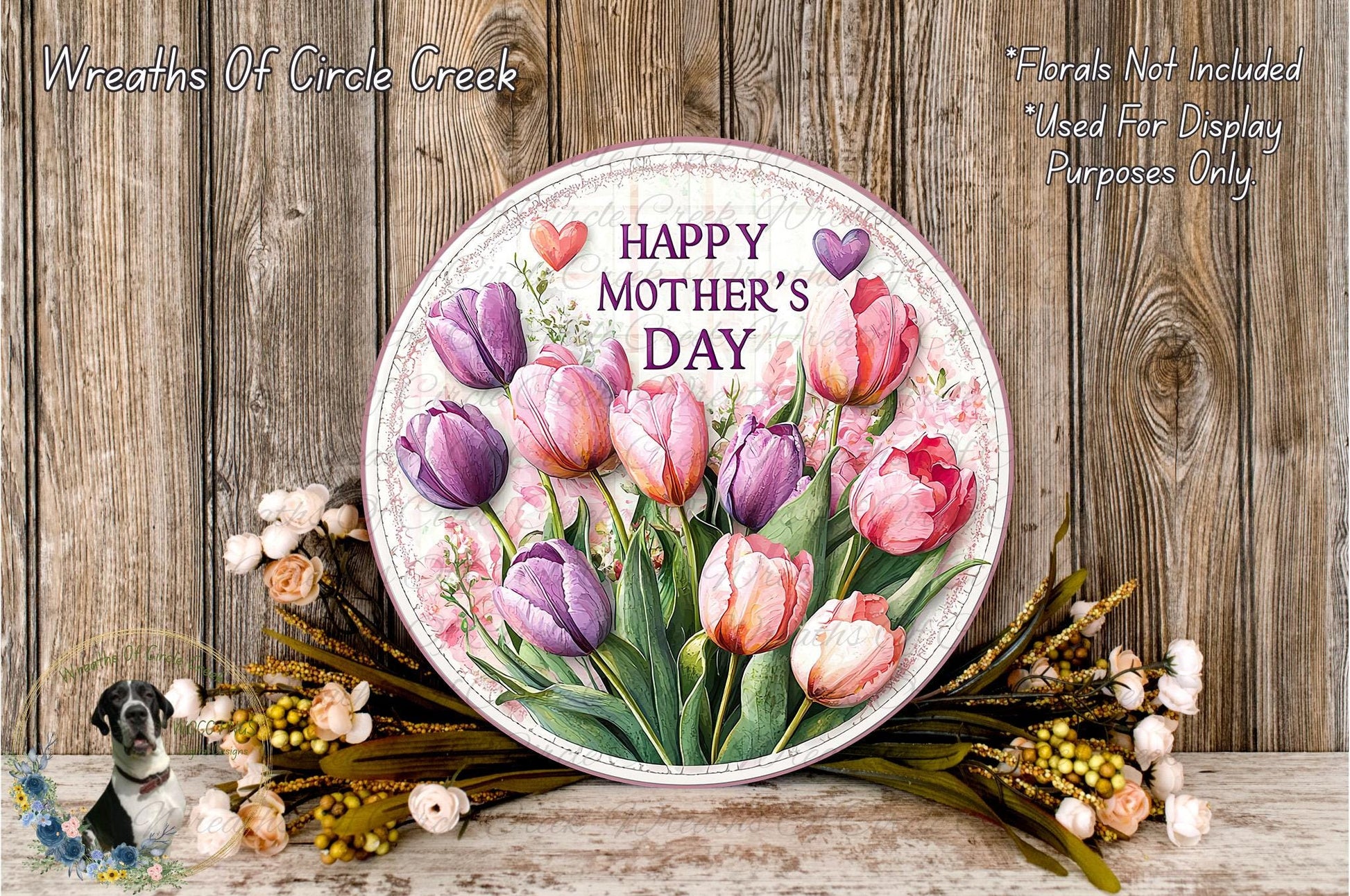 A round floral wreath with a message that reads "Happy Mother's Day" is displayed on a wooden surface, surrounded by various flowers and greenery.