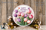 A round floral wreath with a message that reads "Happy Mother's Day" is displayed on a wooden surface, surrounded by various flowers and greenery.