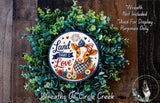 a wreath with a cow wearing a patriotic outfit, surrounded by greenery. The wreath is displayed on a wooden surface, and there is a small dog in the bottom right corner of the image.