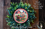 a wreath with a red wagon filled with fruits and vegetables, surrounded by a lush green wreath. The wreath is placed on a wooden surface, and there is a small dog in the bottom right corner of the image.
