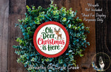 A Christmas wreath made of green leaves and a red and white circular sign that says &quot;Oh Deer, Christmas is Here&quot;.